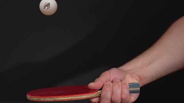 Close-up of hand gripping ping pong paddle firmly. Red and wood colors stand out against black background. Fingers wrapped tightly around the handle. Shows focused grip during practice session