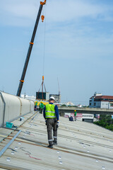 Fototapeta premium A photovoltaic technician wearing a safety vest and hard hat calmly steadies the rigging line of a mobile crane on a large corrugated factory rooftop under the tropical sun in Thailand.