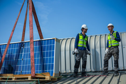 Two renewable energy specialists in hard hats and hi vis workwear steadily discuss panel distribution beside the delivery stack high on an industrial warehouse roof in a Thai city.