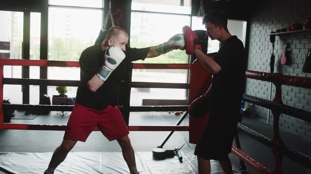 Two men sparring inside boxing ring as one boxer drives straight punches, retreats under pressure, while trainer holds focus mitts in bright backlit gym, with boxing, sparring, trainer, coach detail