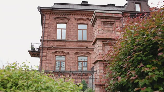 Slow garden-level motion along red brick residential facade with arched windows, small balcony, dentil cornice, and leafy foreground creating layered urban heritage architecture
