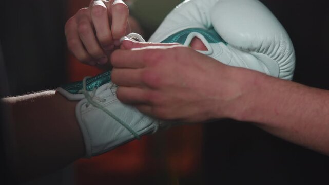Hands tightening frayed lace on white leather boxing glove with teal trim inside dim gym, showing focused preparation, tactile texture, and worn training gear detail