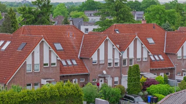 Red brick row houses with orange clay tile roofs in European suburban neighborhood, low angle view of cozy residential homes and green backyard trees in Germany