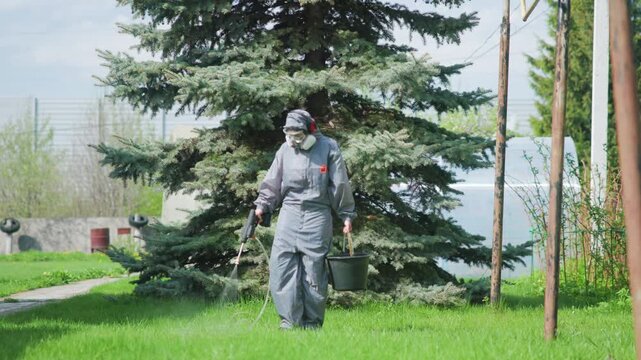 Protective suit worker walking beside evergreen tree while spraying treatment across green lawn in outdoor maintenance setting, with worker, protective, coverall, mask detail