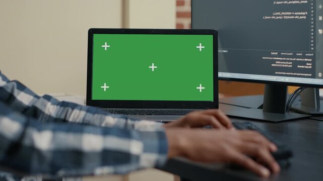 Developer hands type programming code beside a laptop with green screen mockup, creating replaceable screen space for software interface compositing