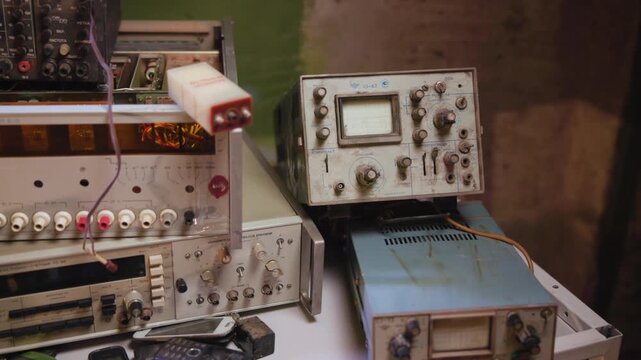 Vintage analog oscilloscopes and test instruments stacked on cluttered workshop bench under warm directional light, with worn cables, knobs, metal housings, and soft shadowed detail