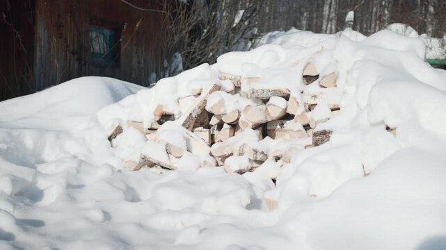 Snow-covered stack of birch firewood beside weathered wooden siding and sparse winter shrubs in quiet residential yard under bright sunlight with crisp shadows, with snow, firewood, birch, logs detail