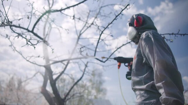 Protective gear worker in hooded coverall and respirator holding sprayer nozzle beside bare orchard branches under soft backlit sky, side profile outdoors, with protective, suit, respirator, mask