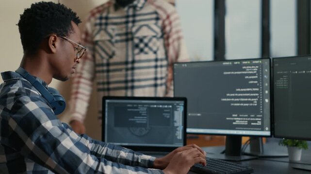 Software developer writes code on a laptop beside multiple computer screens, connecting app development, data parsing and office workflow, during a focused development workflow.