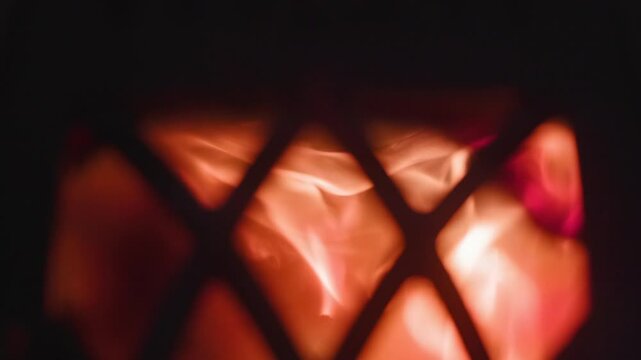 Decorative metal lattice silhouette with warm flames flickering behind diamond grille, creating glowing orange light, deep shadow, textured contrast, cozy atmosphere, and dramatic backlit pattern