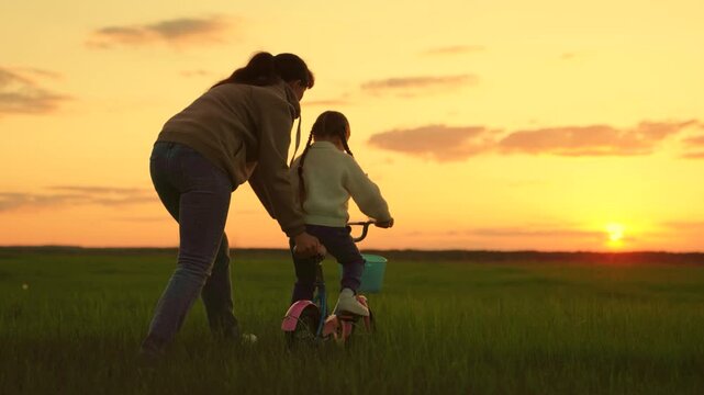 Childhood dream to ride bike. Happy family life mom baby, Parental support. Mom teaches her little daughter to ride bicycle, sunset. Young mother teaches child to keep balance while sitting on bicycle