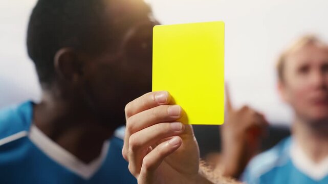 Close focus on yellow card held by referee hand after rules violation during soccer match, discipline due to foul punishment, referee decision with card warning and sanction, blurred stadium.