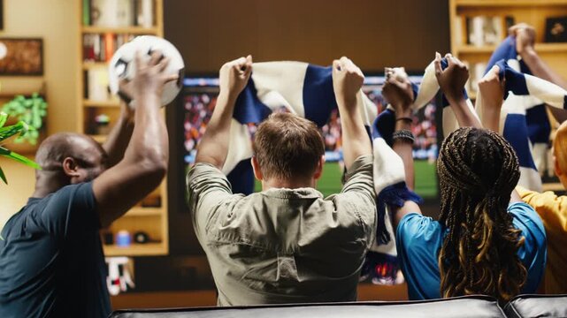 Happy soccer supporters at home clink beer cups after a positive match result as cheering football supporters share an excited couch reaction during live soccer game on TV while drinking alcohol.