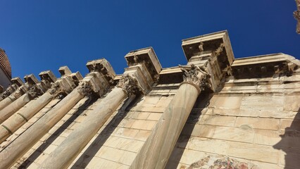 Athens, Greece - 22 June 2026. Low angle view of the western façade of Hadrian's Library with Corinthian columns, carved capitals, entablature and marble wall beneath a clear sky. © Michael
