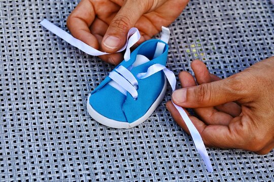 Hands tying the laces on a small blue baby sneaker on a textured surface. A close-up view shows a person's hands carefully tying the white laces on a tiny blue infant shoe