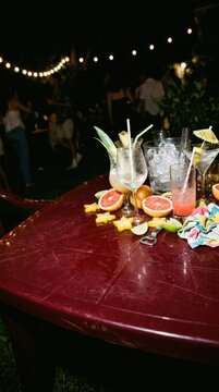 Cocktails and fruit garnish on table at outdoor night party. Tropical drinks with ice bucket served at summer celebration. Festive social gathering with guests in background.