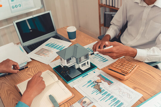Real estate agent explaining housing details and loan conditions to client with house model on desk.