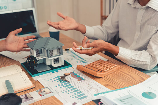 Real estate agent explaining housing details and loan conditions to client with house model on desk.