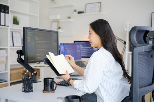 Female programmer writing notes while developing software across multiple computer screens in a modern office.