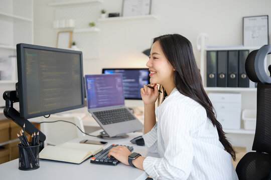 Female programmer writing notes while developing software across multiple computer screens in a modern office.