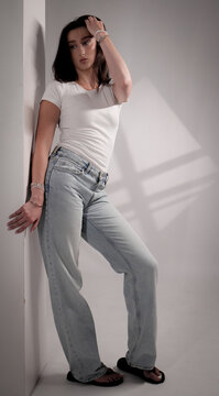 Young woman leaning against a wall in a minimalist studio setting, wearing a white t shirt and jeans