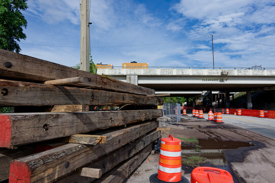 Active road construction under a concrete highway bridge featuring stacked wooden beams, orange traffic barrels, safety barriers and muddy pavement on a bright, partly cloudy day.
