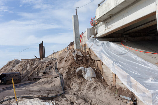 Coastal bridge repair site showing eroded sand embankment, concrete bridge supports, plastic sheeting and construction materials used for stabilization and temporary erosion control during repairs.
