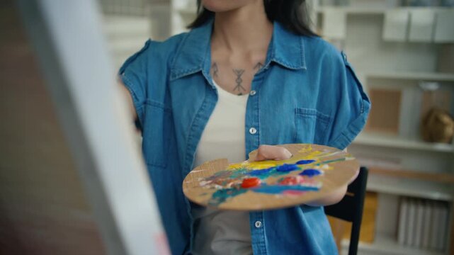 Young Asian woman artist painting on canvas easel with paintbrush in art studio, representing creative professional women designer, fine arts student lifestyle and freelance illustrator
