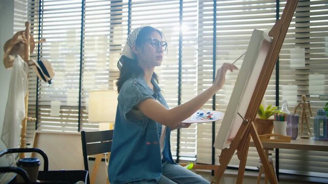 Young Asian woman artist painting on canvas easel with paintbrush in art studio, representing creative professional women designer, fine arts student lifestyle and freelance illustrator