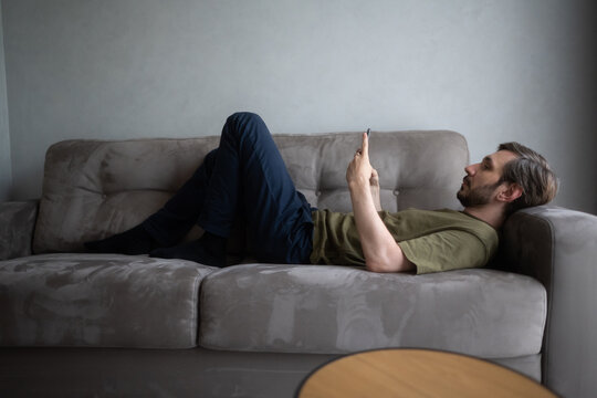 Young man unwinding on a comfortable couch, scrolling and browsing content on his mobile phone, enjoying leisure time and digital connection indoors