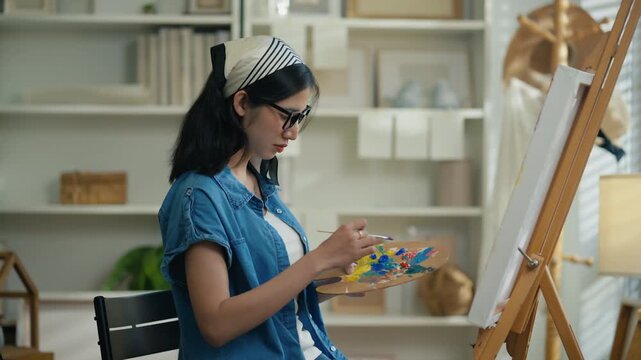 Young Asian woman artist painting on canvas easel with paintbrush in art studio, representing creative professional women designer, fine arts student lifestyle and freelance illustrator