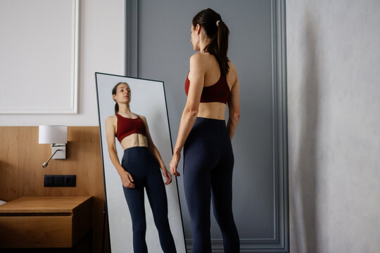Woman standing in sportswear, looking at her reflection in a full length mirror, focusing on her appearance and portraying issues with body image and eating disorders