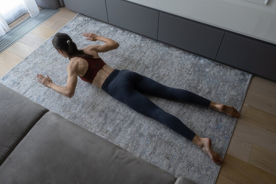Woman lying on a rug in a modern living room, performing a back muscles exercise, focusing on strength training and maintaining a fit and healthy lifestyle with a home workout