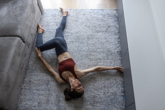 Woman performing a supine spinal twist, stretching her body on a rug by a sofa, engaging in a yoga flow for flexibility, relaxation, and physical wellbeing at home