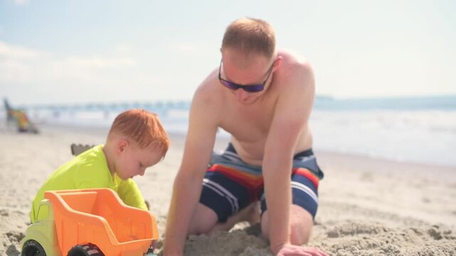 Caucasian father and young boy engage in playful activities on a sandy beach, using a colorful toy truck and shovel while digging and exploring the sand under bright sunlight and ocean waves