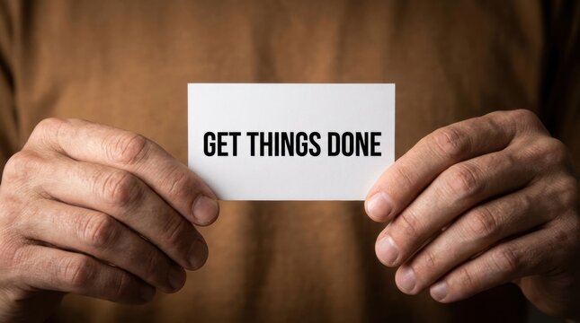 Close up of human hands holding a white paper card displaying the motivational text get things done against a background of brown