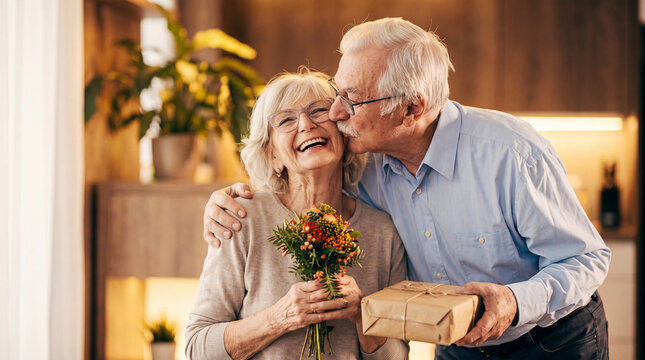 A loving senior man kisses his smiling wife on the cheek while she holds a bouquet of flowers and a gift, celebrating a special occasion at home.