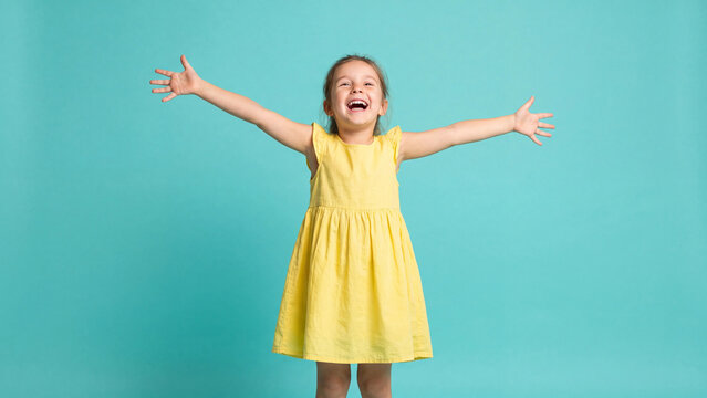 Girl standing with arms spread wide in a bright yellow dress against a solid turquoise background expresses pure joy and excitement during a fun studio photoshoot session.