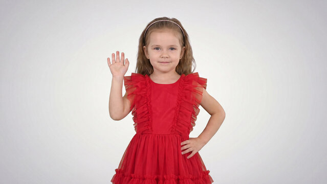 Girl in a red dress stands against a light background while waving her hand to welcome visitors into the studio setting today.