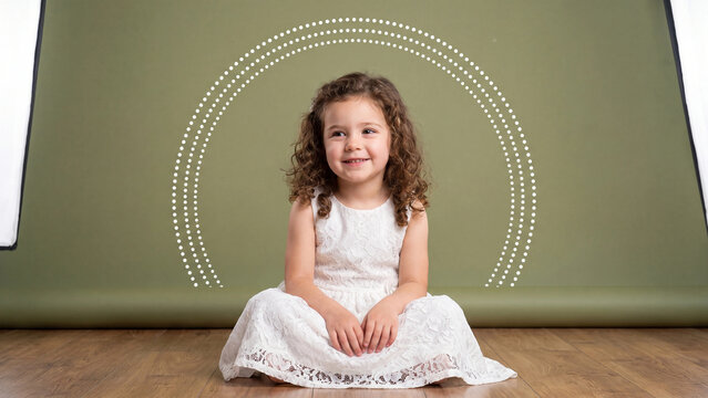 Girl with curly hair sits on a wooden floor wearing a white dress in front of a green studio background with a dotted rainbow design.