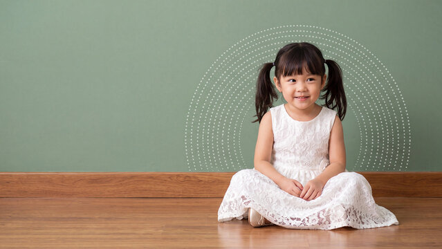 Toddler with pigtails wearing a white lace dress sits on a wooden floor in front of a green wall featuring a white dotted rainbow.
