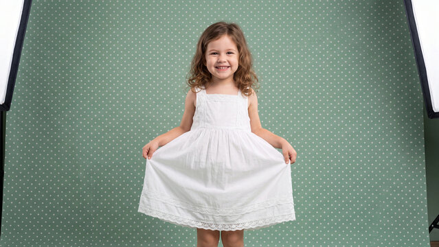 Child standing in a white dress smiles brightly against a green patterned background while illuminated by professional studio lighting.