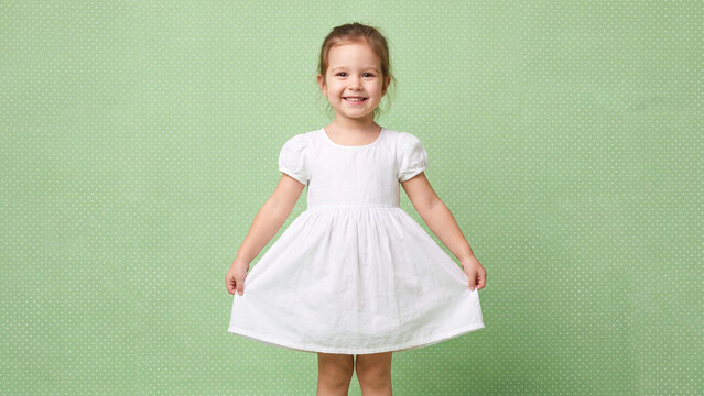 Girl wearing a simple white dress poses joyfully while holding the fabric of her skirt against a solid green polka dot textured wall surface.
