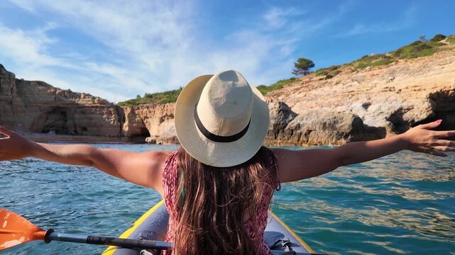Woman in a kayak celebrating freedom with open arms while paddling towards a sunny beach with beautiful sea caves. Outdoor adventure and summer travel vacation exploring the stunning coastline