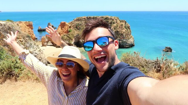 Joyful young couple taking a selfie video while on vacation, smiling and laughing together with a beautiful scenic view of the cliffs and turquoise ocean in the background