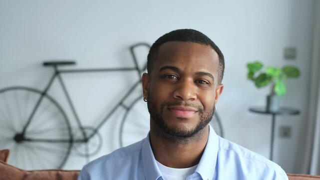 Close-up portrait of a young, confident, handsome african american man looking at the camera and smiling, sitting on a sofa in his modern living room. Happy millennial guy posing at home