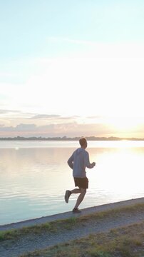 Vertical Drone tracking shot from behind of a man running along a riverbank at sunset. Male athlete jogging on a levee during outdoor cardio fitness training for an active lifestyle