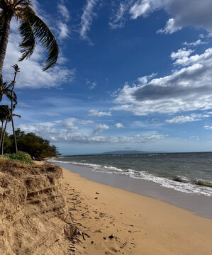 Scenic tropical coastline at Waipuilani Beach in Kihei, Maui, Hawaii, with palm trees, ocean waves, sandy shoreline, dramatic clouds, and expansive blue sky.