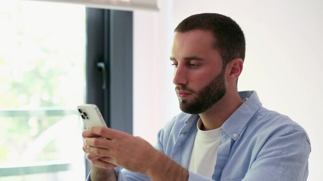 Handsome caucasian man with a beard sitting by the window, using his mobile phone to browse social media, smiling and typing messages while enjoying a relaxing moment in a bright apartment