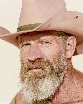 Rugged bearded man in a weathered cowboy hat with a steady gaze and sun-kissed skin against a soft cream background for lifestyle and western themes